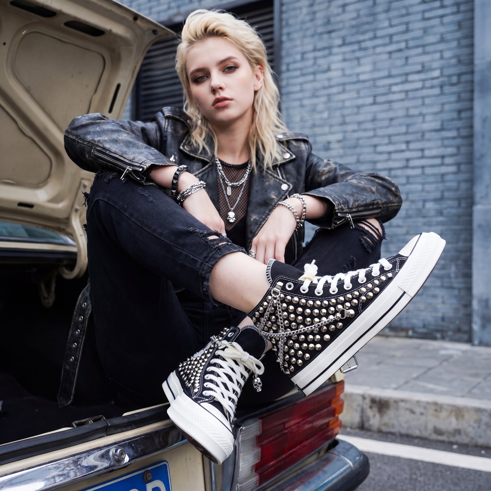 Young female model in punk street style sitting on an open car trunk wearing black studded Converse sneakers with chains