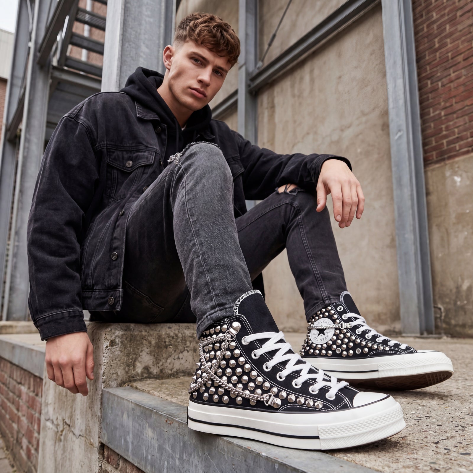 Young male model in black denim and hoodie sitting on concrete steps wearing black studded Converse sneakers with chains